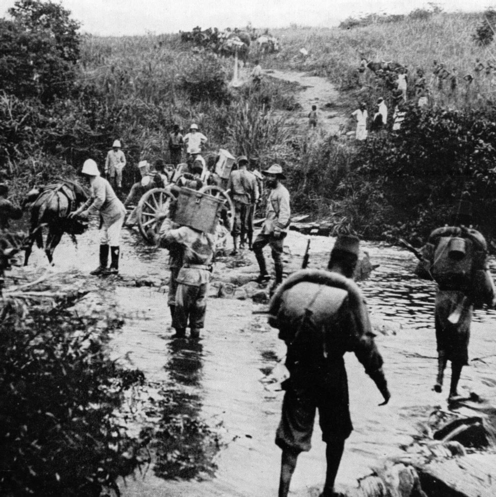 Photographie historique en noir et blanc de la Force Publique du Congo Belge en campagne, 1918. Des soldats congolais en uniforme, portant des armes et des équipements, marchent en colonne dans un paysage africain. Ils sont les héros oubliés de l'histoire mondiale.
