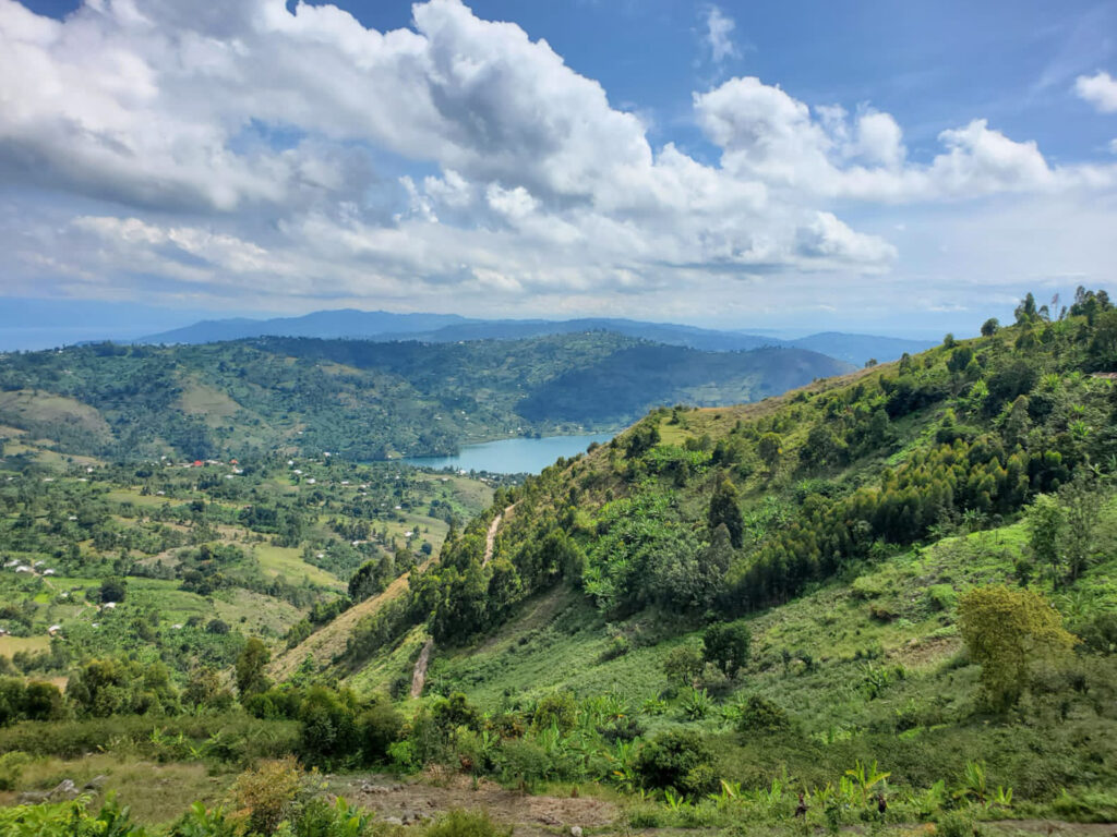 Panoramic view of a green valley with a blue lake, scattered houses, and rolling hills under a partly cloudy sky.