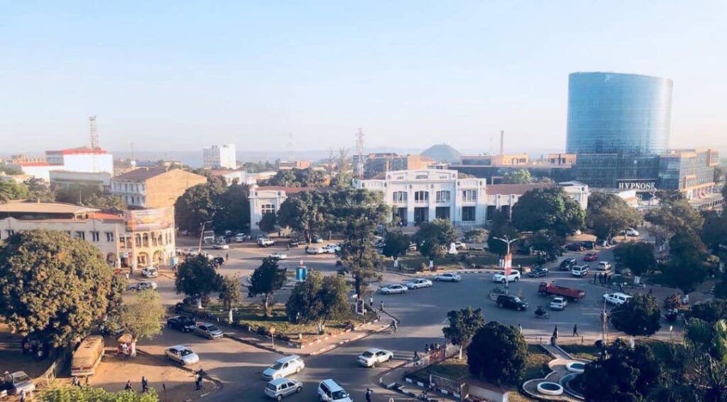 Aerial view of a city square with a roundabout, trees, white colonial buildings, and a tall cylindrical glass building on the right.