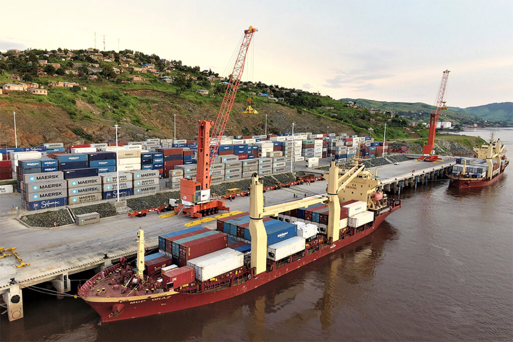 Container ship docked at a port with stacked cargo containers and tall cranes, hillside town in the background.